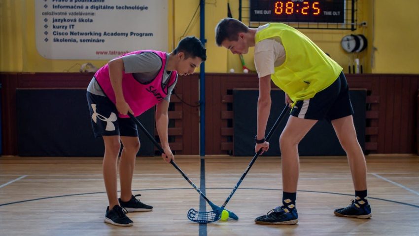 Two teenagers face off in a floorball match inside a sports gym with equipment visible in the background.