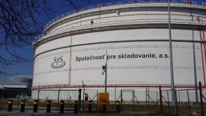 Wide landscape photo of large oil storage tanks at the Bučany petroleum storage facility in Slovakia, set behind a green field under a clear, pale blue sky.