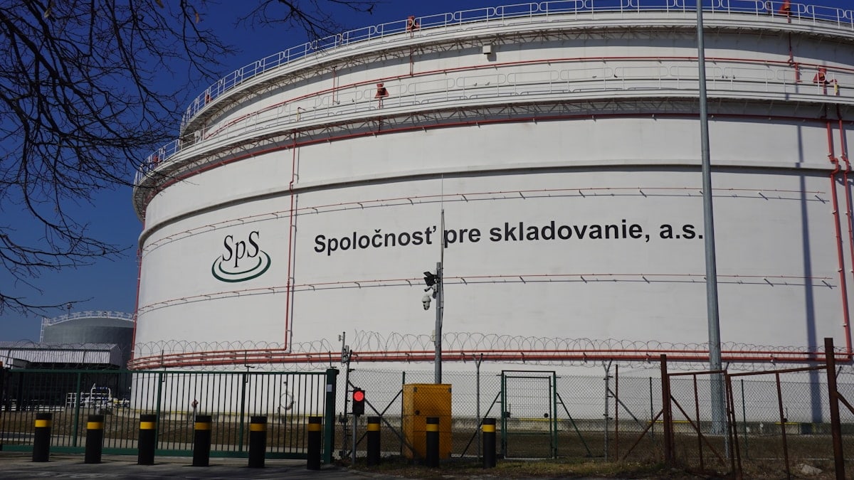 Wide landscape photo of large oil storage tanks at the Bučany petroleum storage facility in Slovakia, set behind a green field under a clear, pale blue sky.